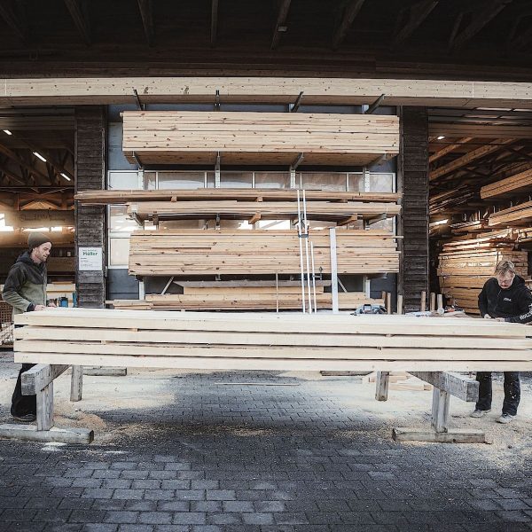 Zwei Personen arbeiten in einem Sägewerk oder Holzlager mit Stapeln von Holzbrettern, umgeben von Regalen mit Holz.
