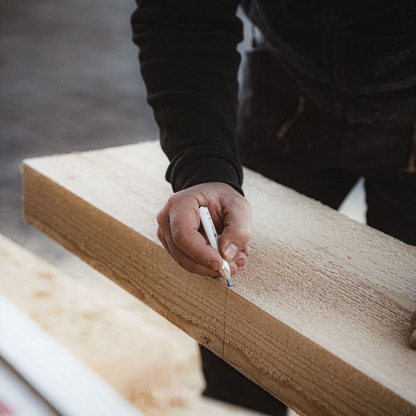 Eine Person markiert mit einem Bleistift eine gerade Linie auf einem Holzbrett und bereitet das Holz zum Schneiden vor.