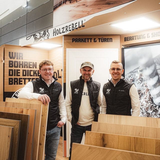 Drei Männer stehen in einem Ausstellungsraum mit Mustern von Holzfußböden, und an den Wänden hinter ihnen sind Schilder in deutscher Sprache über Parkett, Türen und Beratungsdienste zu sehen.